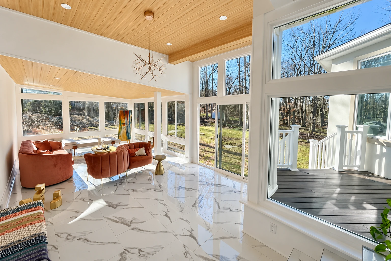 Bright sunroom with wood-paneled vaulted ceiling, marble-look floor, rust-colored chairs, and large windows overlooking a wooded yard.