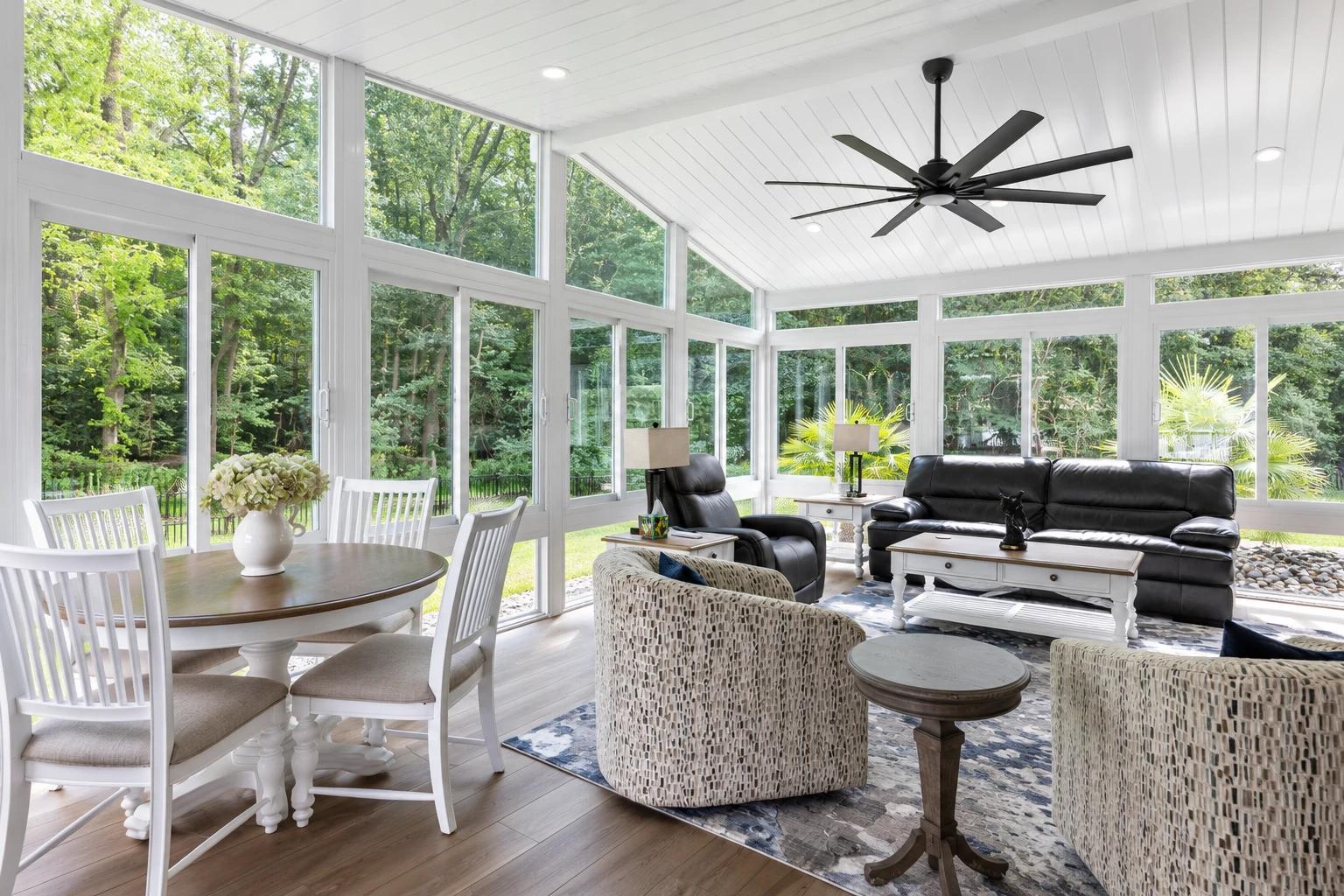 Bright three-season sunroom with expansive windows, a dining table, sofas and a ceiling fan.